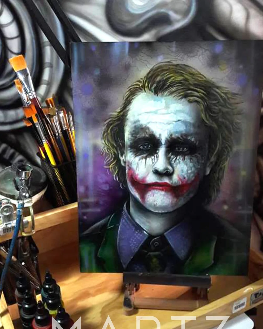 A painted Joker portrait with a white face, dark eyes, green hair and a red smile on a canvas easel.