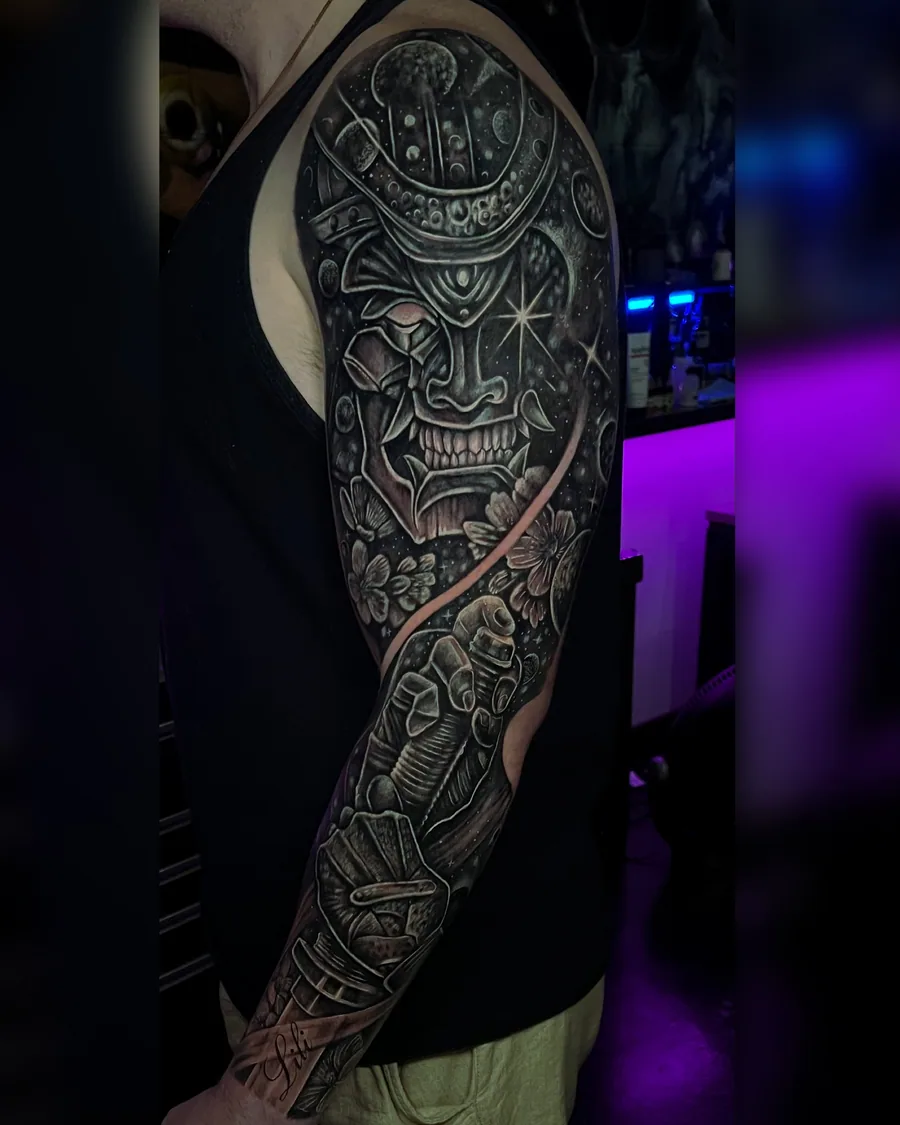 A black-and-gray full-arm tattoo with a samurai mask, celestial armor, blossoms, stars and an armored hand.