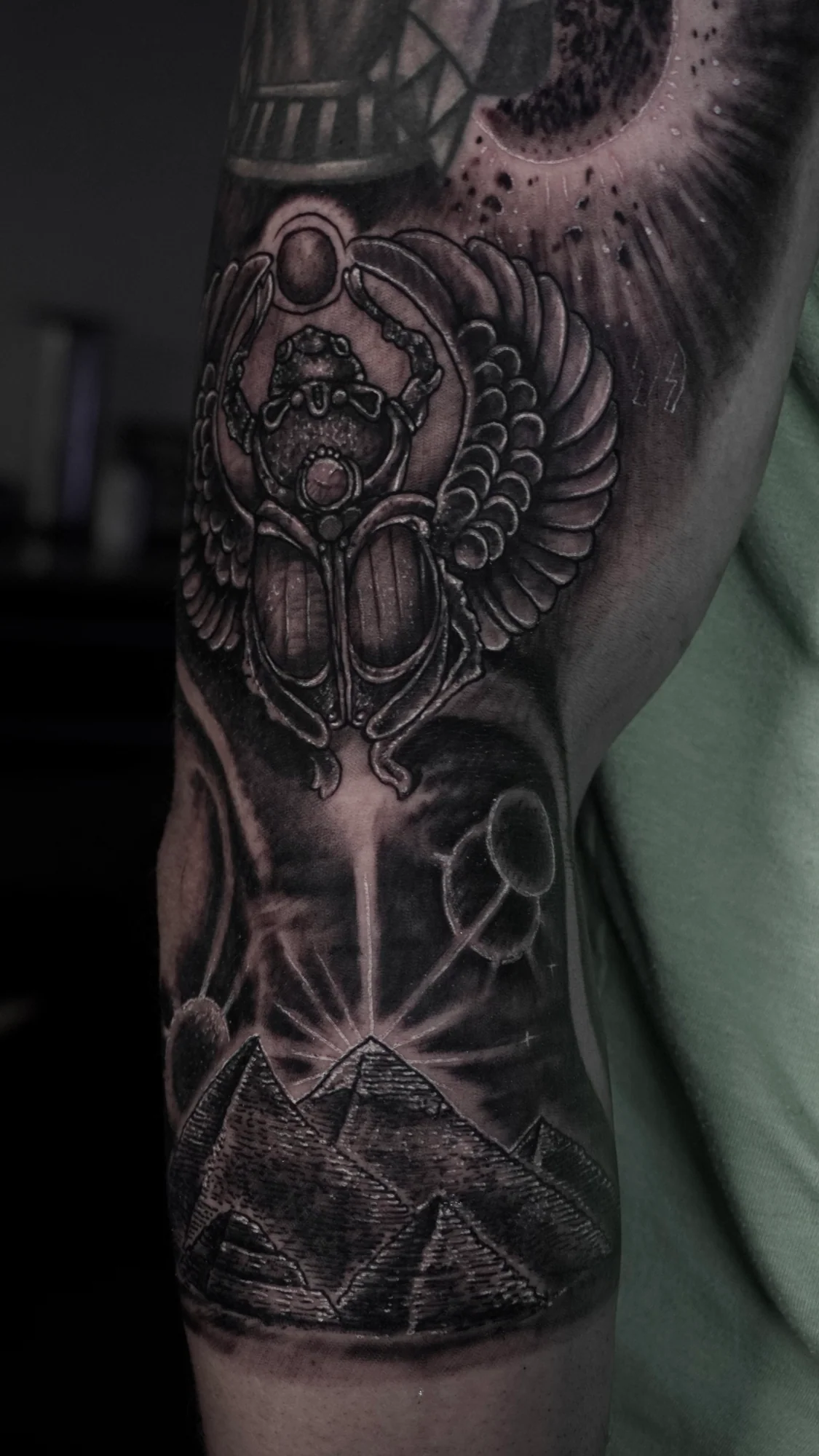 A black-and-gray arm tattoo combining a winged scarab, celestial forms, pyramids and symmetrical ornamental detail.
