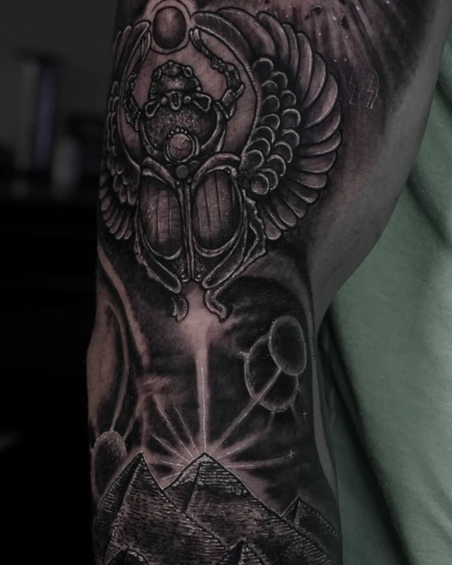 A black-and-gray arm tattoo combining a winged scarab, celestial forms, pyramids and symmetrical ornamental detail.