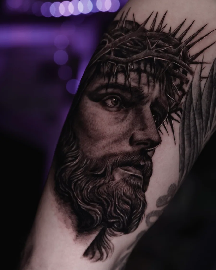 A black-and-gray realistic portrait tattoo of Jesus wearing a crown of thorns.