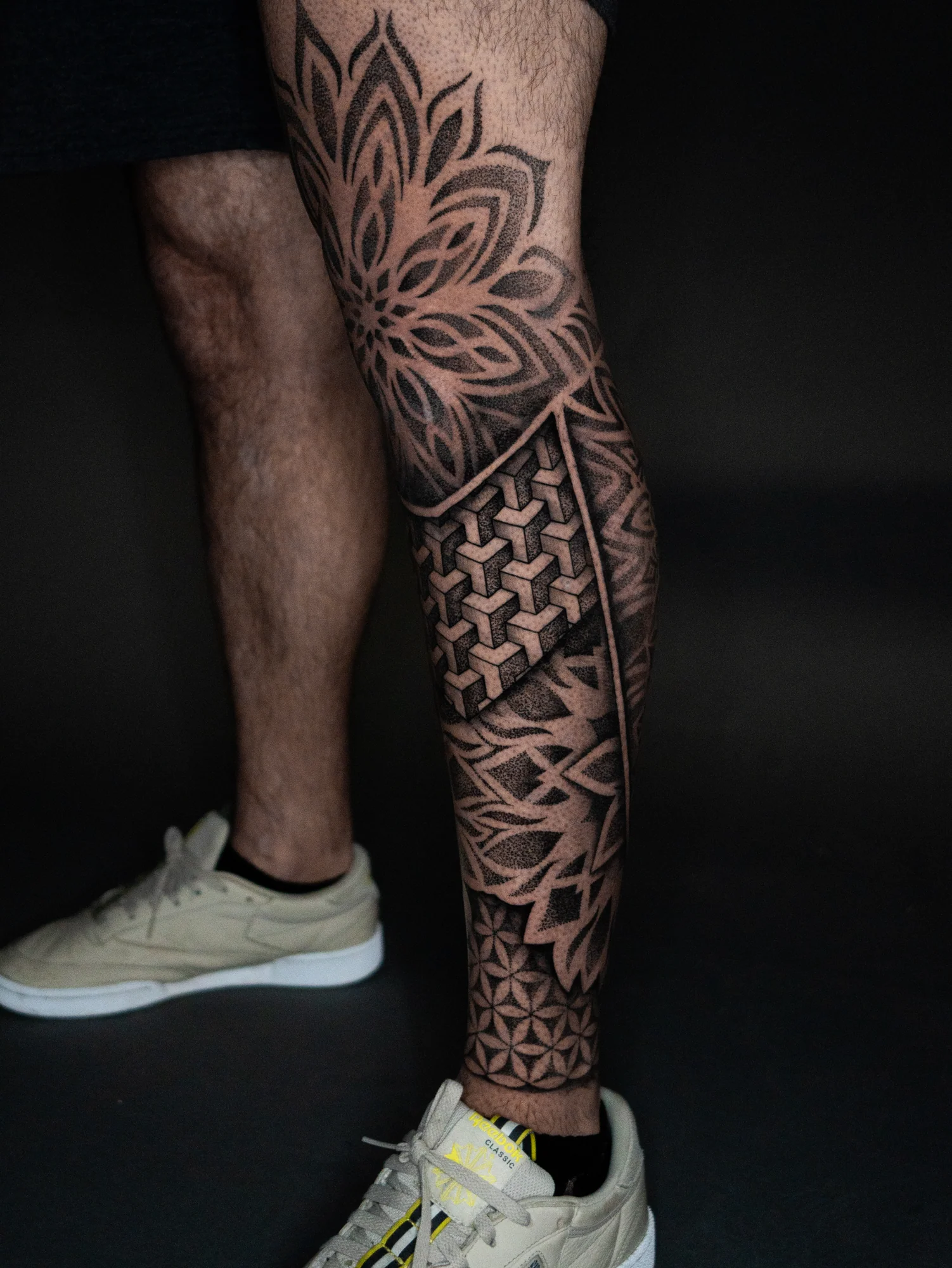 A black-and-gray geometric leg tattoo combining dotwork petals, interlocking cubes and repeating star patterns.