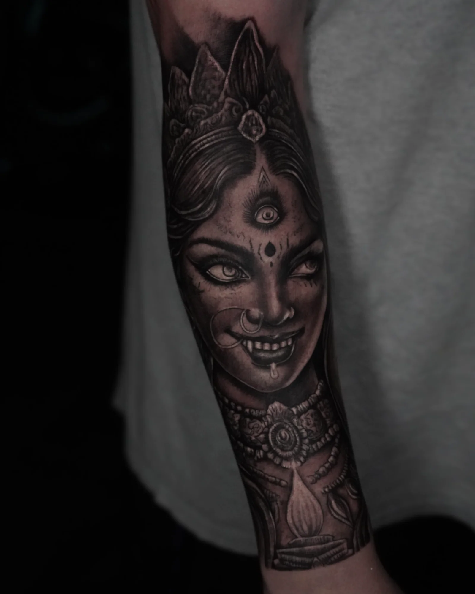 A black-and-gray portrait tattoo of a crowned three-eyed sacred figure with facial jewelry and an ornamental necklace.