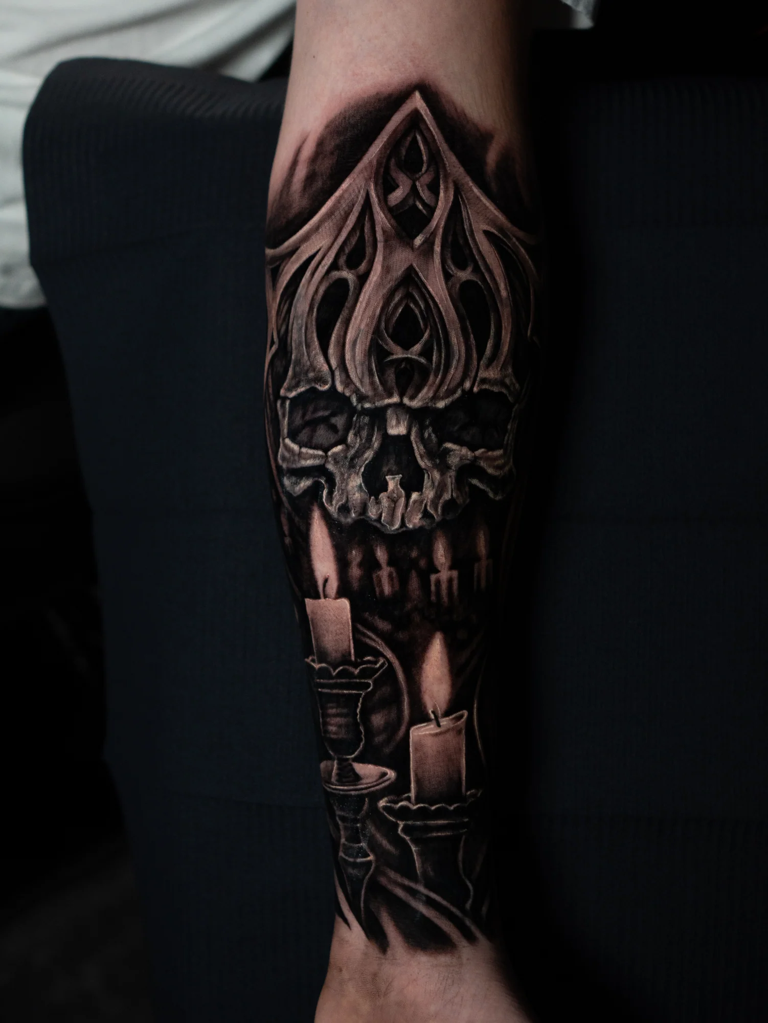 A black-and-gray calf tattoo of a skull beneath pointed Gothic ornament with glowing candles below.