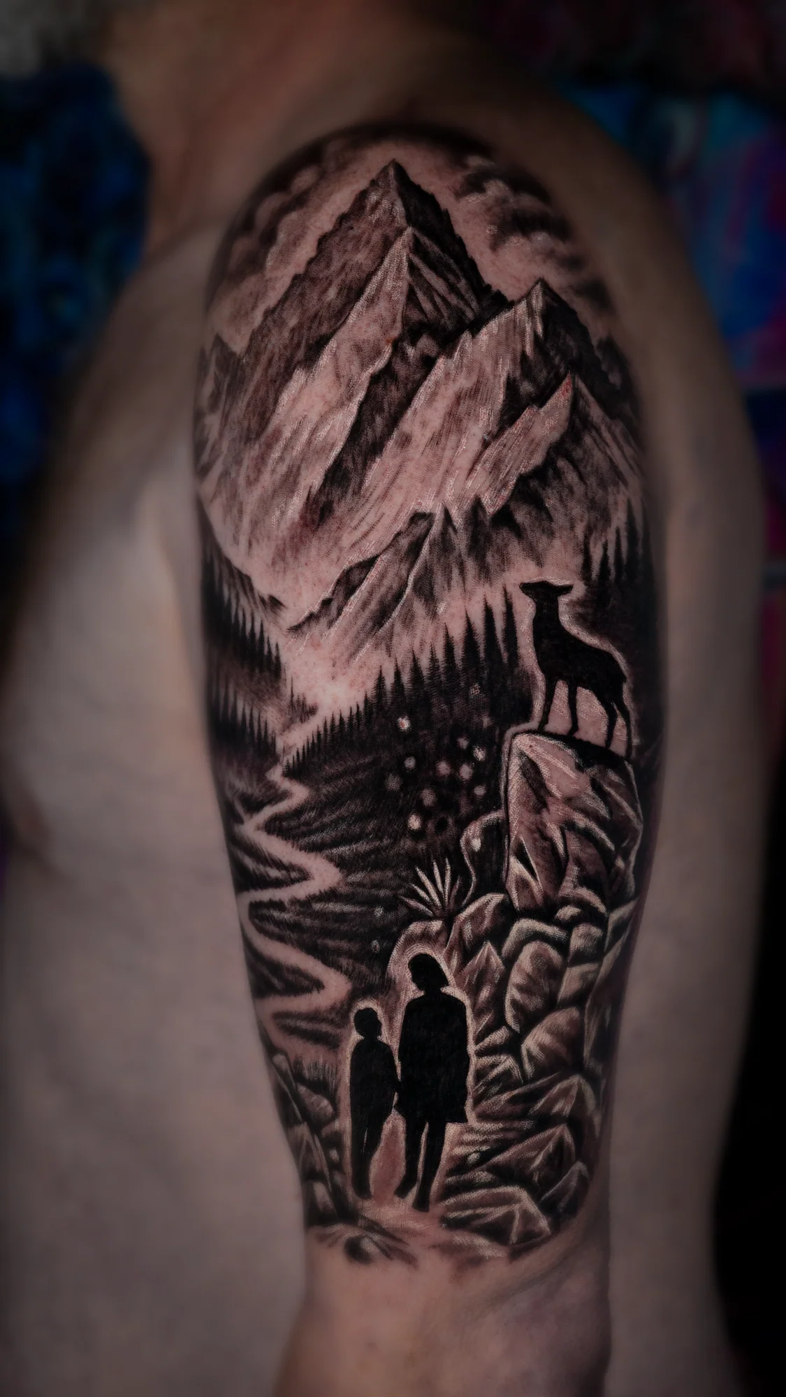 A black-and-gray mountain landscape tattoo with pine forest, a deer on a rock and adult-and-child silhouettes walking together.