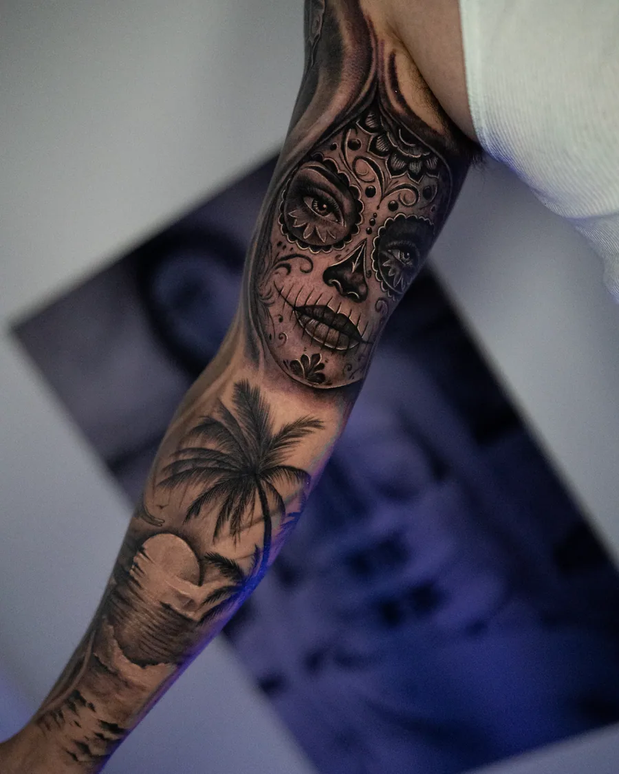 A black-and-gray sleeve combining a decorative sugar-skull portrait with palm trees and a tropical sunset.