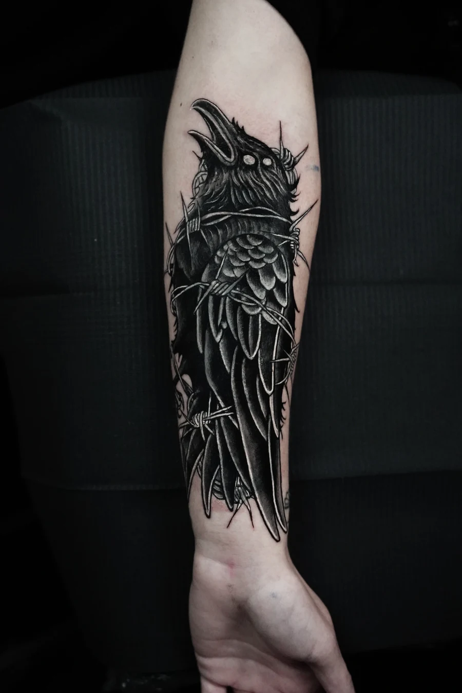 A finished black-and-gray raven cover-up with layered feathers and barbed wire on the forearm.