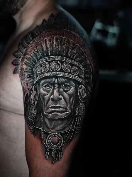 A black-and-gray upper-arm portrait of a figure wearing a detailed feathered headdress.