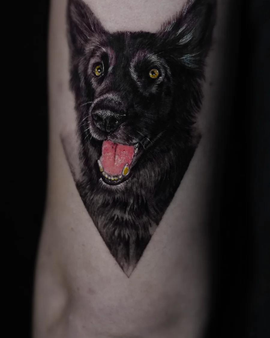 A portrait tattoo of a dark-coated dog with upright ears, bright eyes, and its tongue showing.