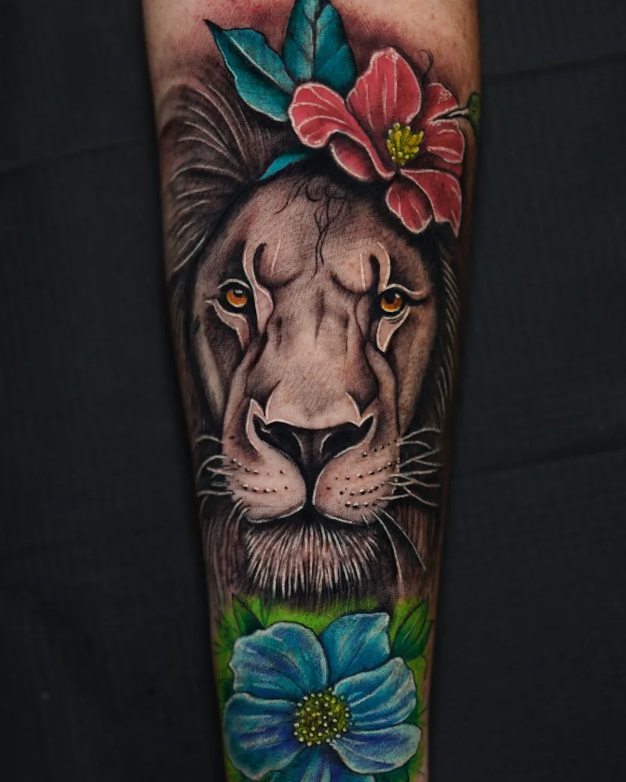 A lion portrait framed by red and blue flowers and green leaves in a vertical color tattoo.