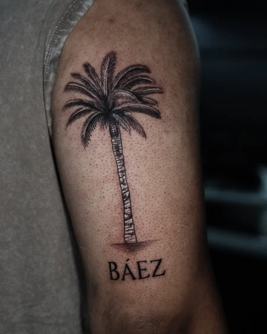 A finely shaded palm tree tattoo with small lettering beneath its trunk on the upper arm.