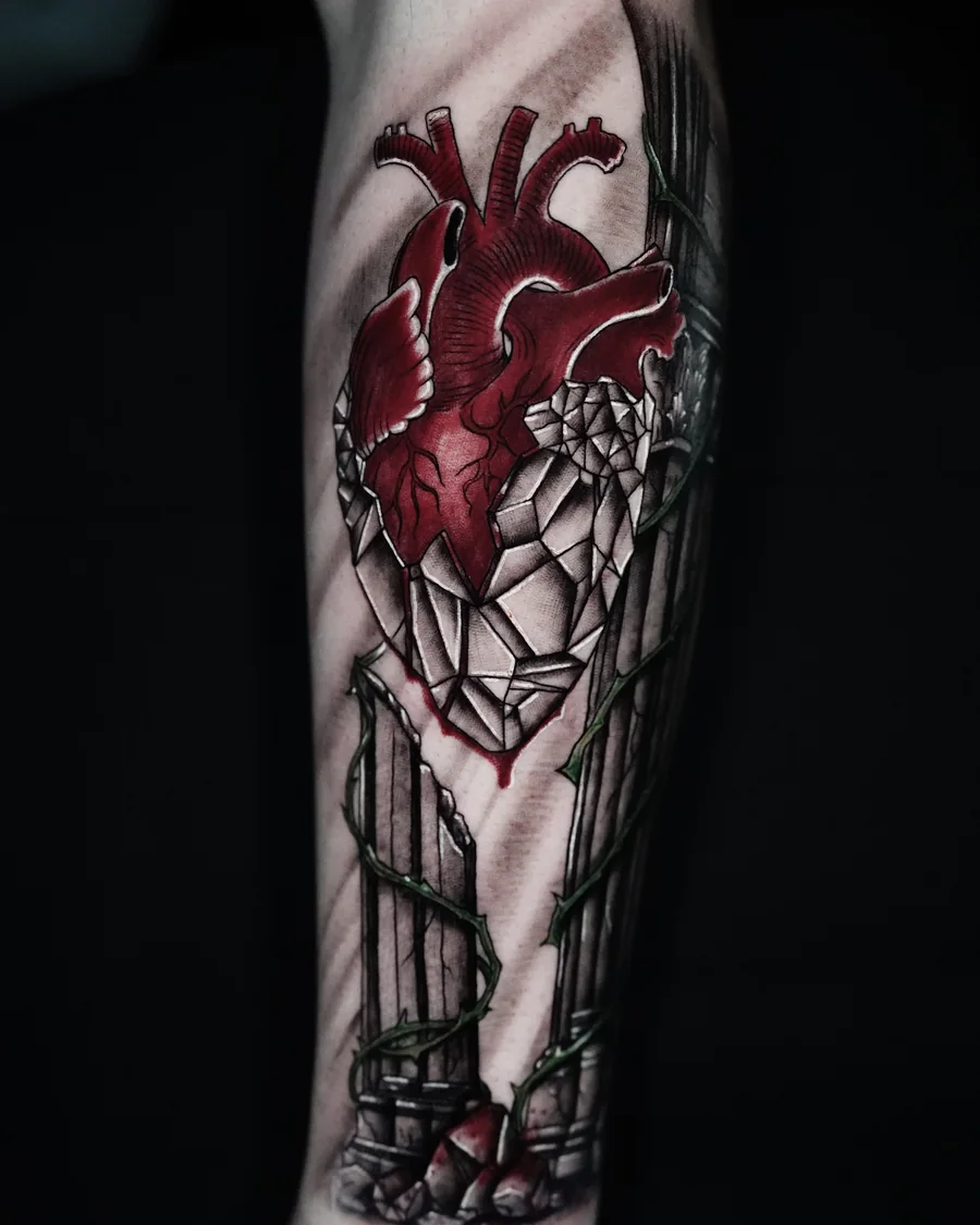 A red anatomical heart above a pointed stained-glass-inspired window in a vertical forearm tattoo.