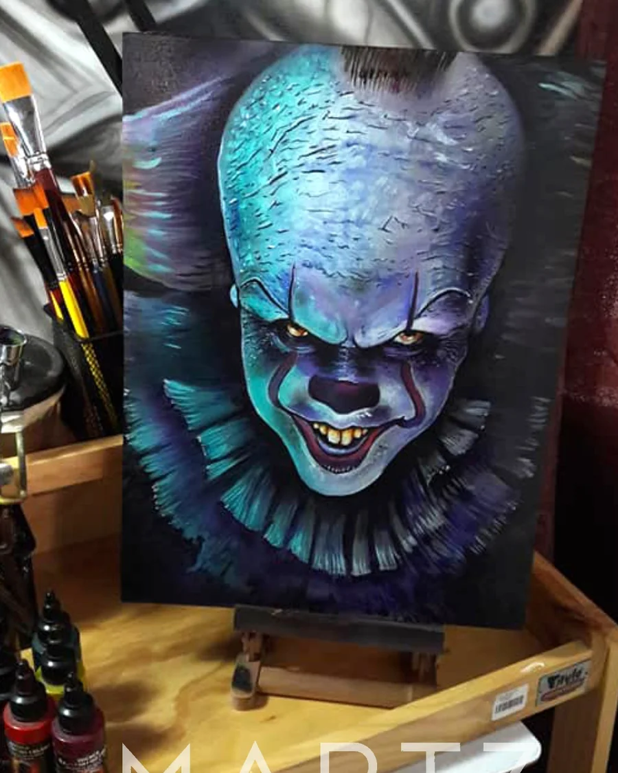 A painted Pennywise clown portrait with a pale blue forehead, yellow eyes and dark violet shadows on a canvas easel.
